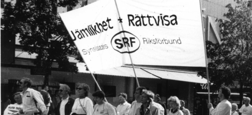 Svartvitt foto demonstrationståg på gata. Stor banderoll med text "Jämlikhet + Rättvisa" Synskadades Riksförbund SRF.