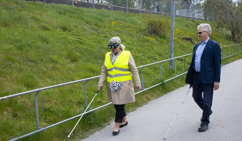 Camilla och Niklas går bredvid varandra på en gångbana. De går båda med vita käppar. Ministern har även gul varselväst med texten "Synskadad" och fingerad optik.