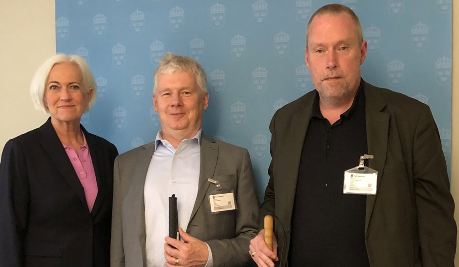 Acko, Niklas och Jimmy står bredvid varandra med en blå vägg med Regeringskansliets logga bakom sig. De har alla tre mörka kavajer och ler litegrann. Niklas och Jimmy håller i sina vita käppar.