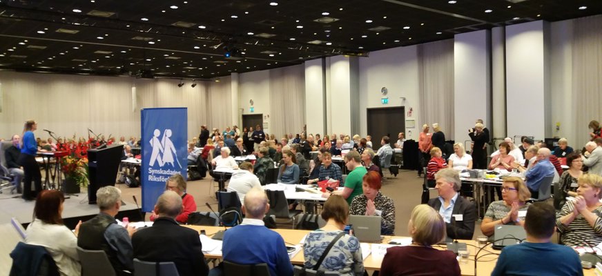 Stor konferenslokal, mycket bord och stolar, människor. Roll up med SRF:s blå färg och logotyp.