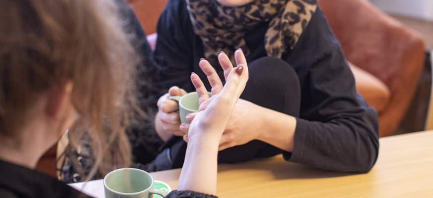 Två personer sitter mittemot varandra vid ett bord och pratar engagerat över en kopp kaffe.
