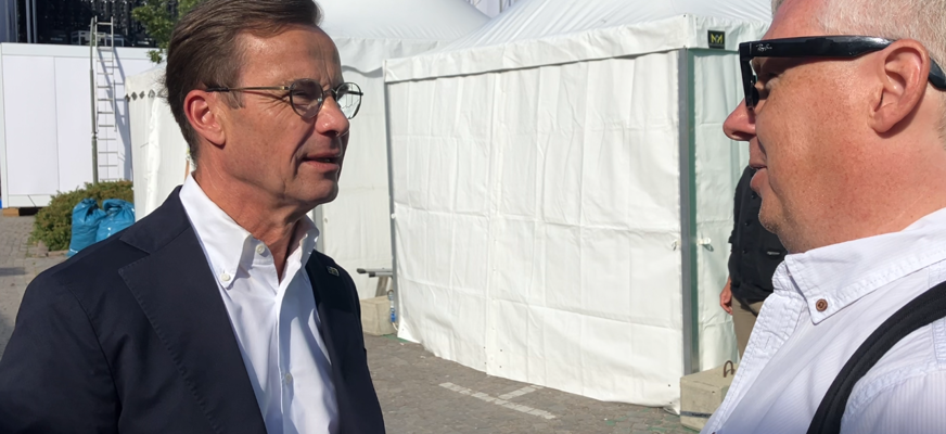 Niklas Mattsson intervjuar statsminister Ulf Kristersson utanför några tält i Visby. Ulf är klädd i svart kostym och vit skjorta. Han har glasögon och bakåtkammat hår. Niklas är klädd i vit skjorta och har solglasögon.