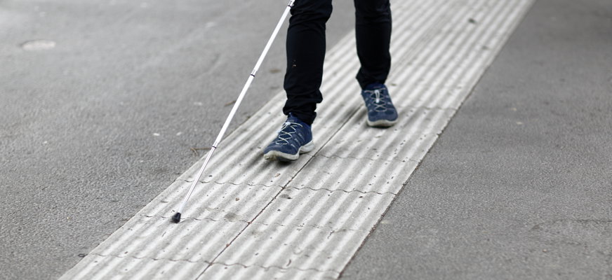 Benen på en person som promenerar med vit käpp på ett ledstråk, omgivet av asfalt.