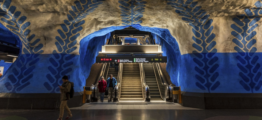 En tunnelbanenedgång där två rulltrappor finns centrerade på bilden, emellan dem finns en trappa. En man och kvinna kliver av den vänstra rulltrappan. Tunnelbanestationens väggar är blå- och vitmålade med mönster i en mörkare blå nyans.