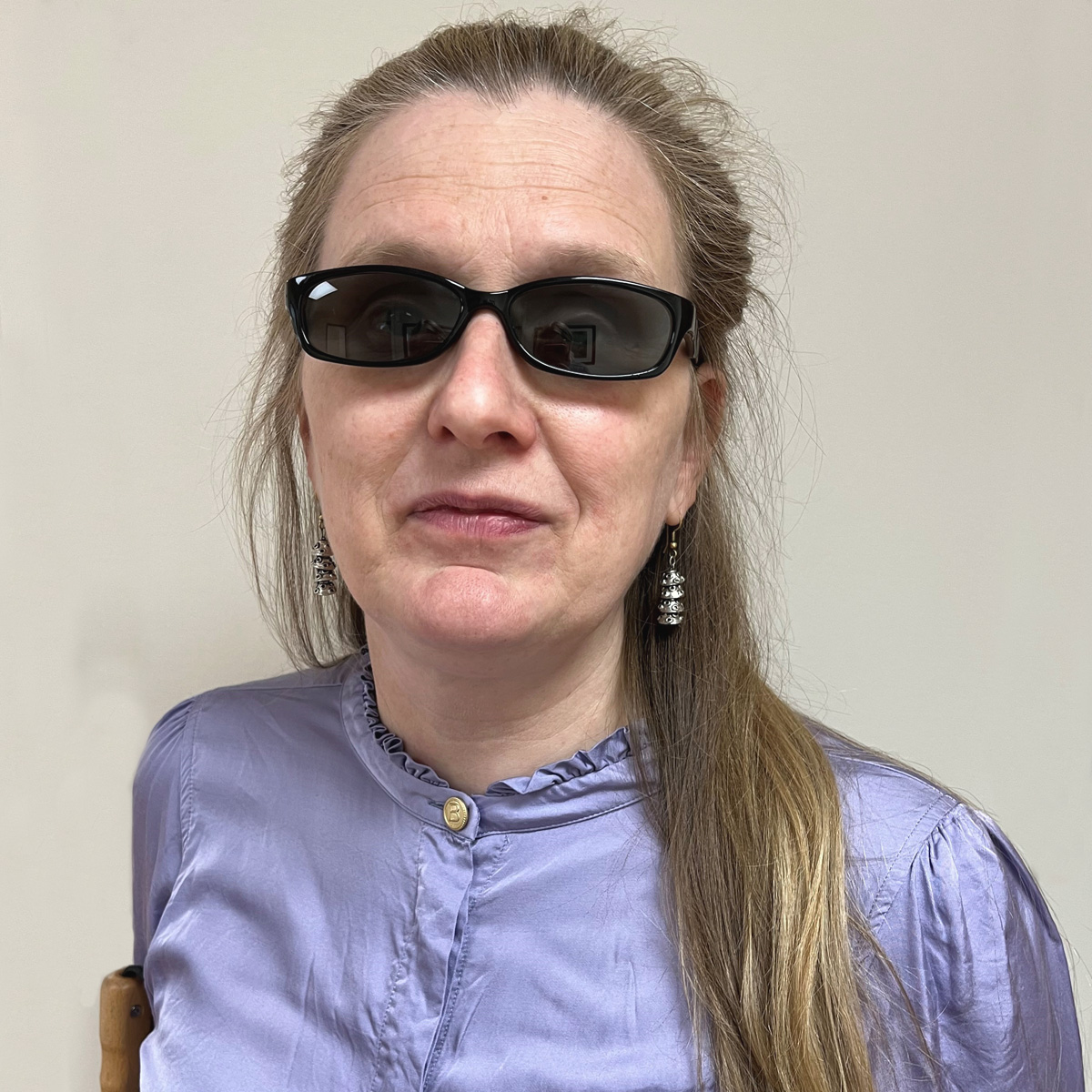 Kvinna i 45-årsåldern, långt rågblont hår i hästsvans, ljuslila sidenblus, silverörhängen, svarta solglasögon.