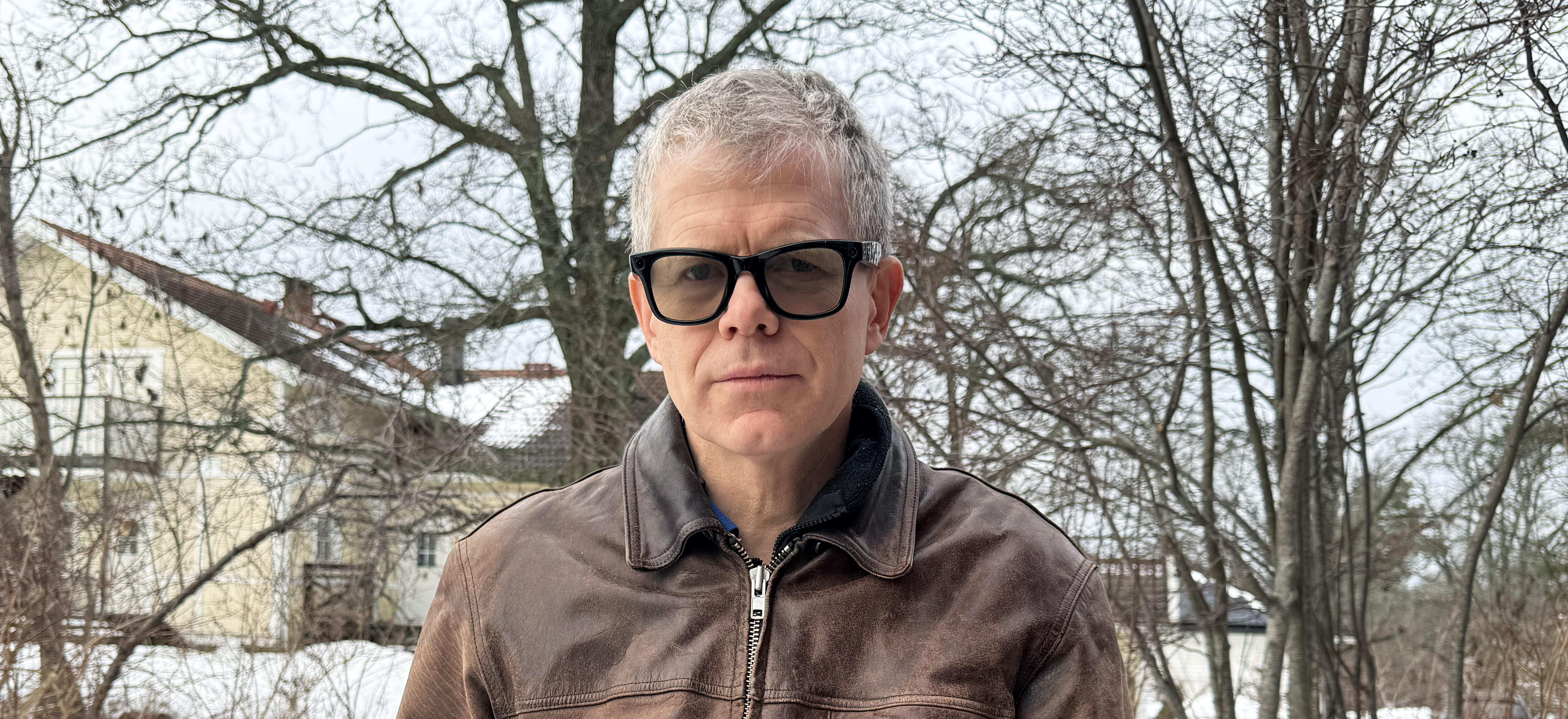 Porträttbild på Henrik Rüffel, en man i kort grått hår och glasögon med tjocka, svarta bågar. Henrik är klädd i en brun skinnjacka och står ute i grått och snöigt landskap. I bakgrunden syns ett gult hus och kala träd.
