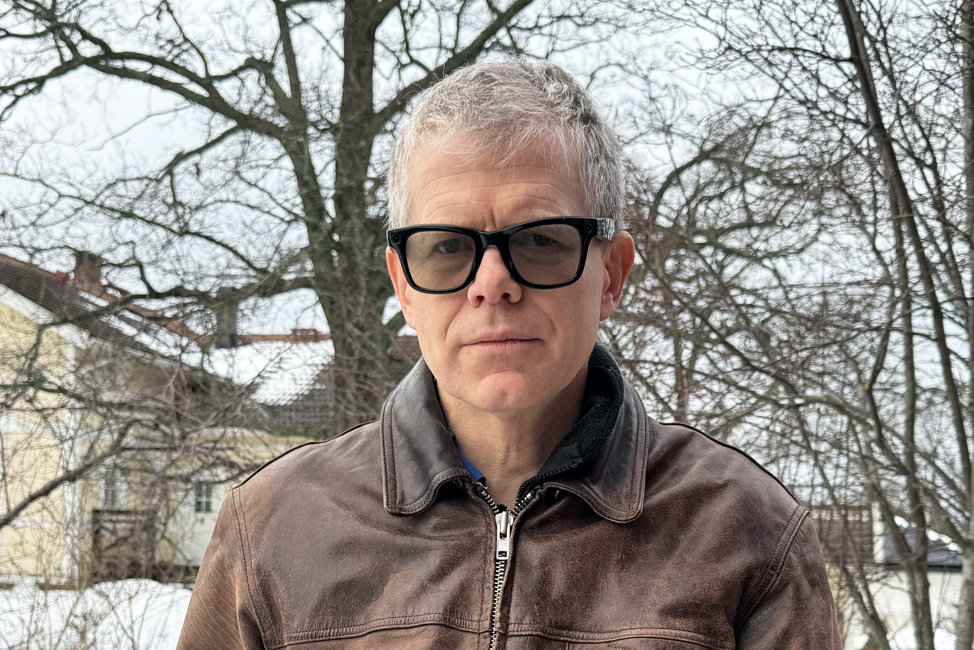 Porträttbild på Henrik Rüffel, en man i kort grått hår och glasögon med tjocka, svarta bågar. Henrik är klädd i en brun skinnjacka och står ute i grått och snöigt landskap. I bakgrunden syns ett gult hus och kala träd.