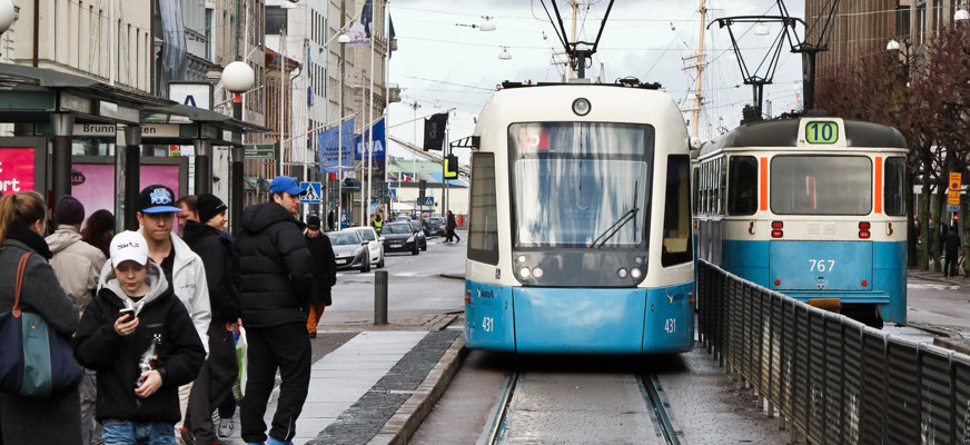 En blå spårvagn bromsar in vid en hållplats i Göteborg. Människor i höstjackor väntar vid hållplatsen, runtom syn flervåningshus och butiker.
