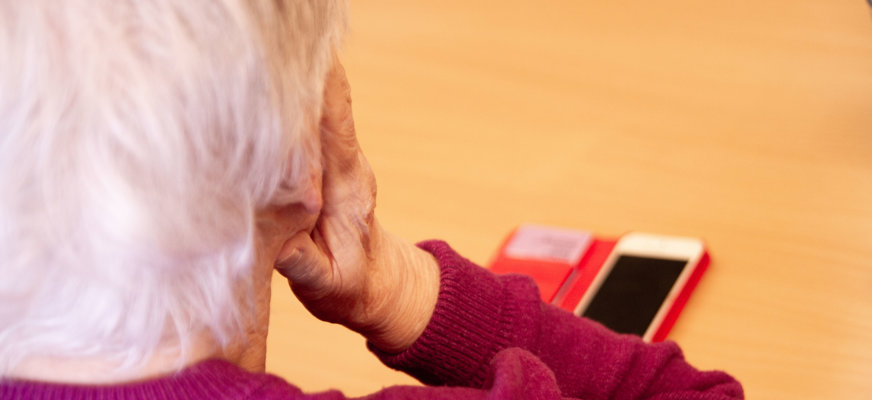 Äldre dam sedd snett bakifrån. Hon lutar huvudet i händerna. Framför henne på bordet syns en mobiltelefon.