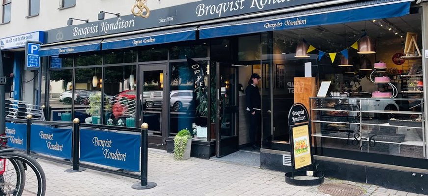 Bilden visar Broqvist Konditori från gatan. Man ser fina bakverk i skyltfönstret samt ser man uteserveringen som har ett blått staket runt borden.