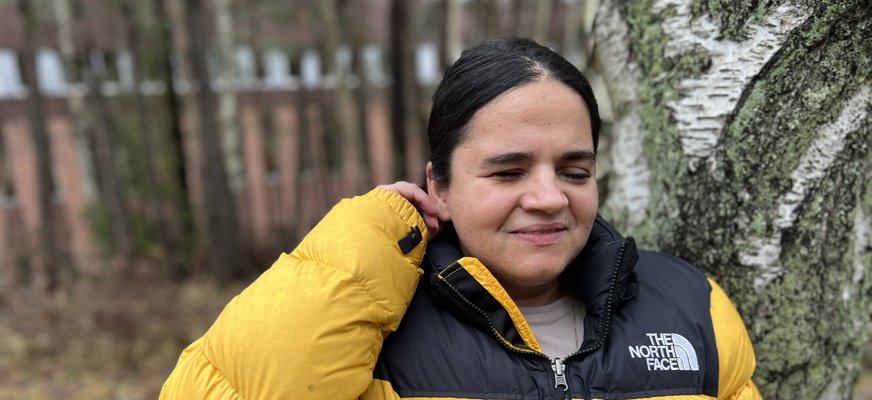 Porträttfoto Sandra, svart hår i hästsvans, knallgul täckjacka lutar sig mot björkstam.