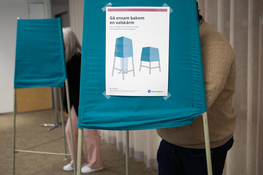 En röstlokal med flera blå valbås. På det främsta båset sitter en skylt med rubriken ”Gå ensam bakom en valskärm”. En person står bakom ett av båsen.