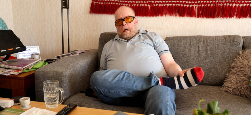 Benny Rosdahl sitter bakåtlutad i en soffa. Han har kort skägg och bär glasögon med orangefärgat glas och en kortärmad pikétröja.