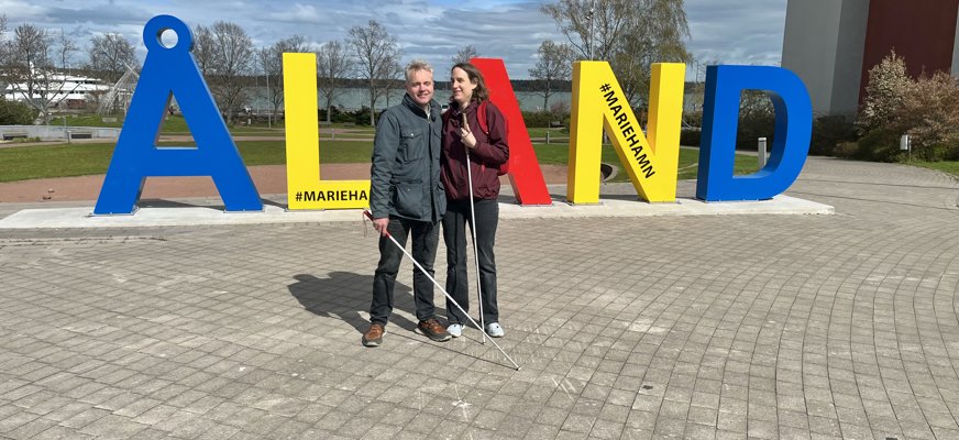 En man och en kvinna står och håller om varandra. De har varsin vit käpp. Bakom dem står bokstäverna Å L A N D som tillsammans bildar ett gult, rött och blått monument. 