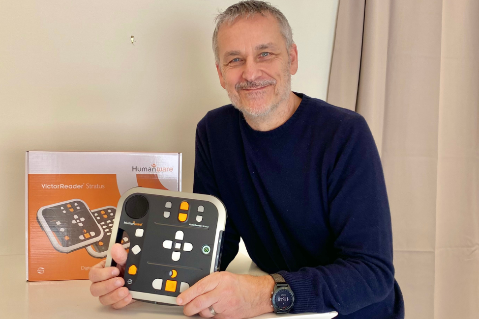 En vit medelålders man med kort grått hår, grå skäggstubb och mörkblå stickad tröja håller i en Daisy bandspelare. Vit vägg bakom.