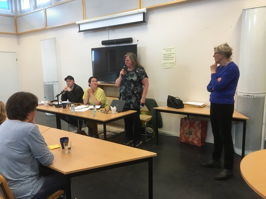 SRF Uppsala läns ordförande Tomas Wallén sitter bredvid årsmötets ordförande, riksdagsledamot Lina Nordquist (L) och lyssnar på årsmötets sekreterare Gunilla Thomsson som står och pratar i en mikrofon. Gruppledsagare Lotta Granér står en bit bort och tittar på.