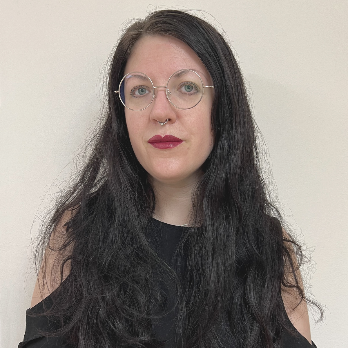 Kvinna i 40-årsåldern, långt svartfärgat hår, runda glasögon, näspiercing, mörkrött läppstift.