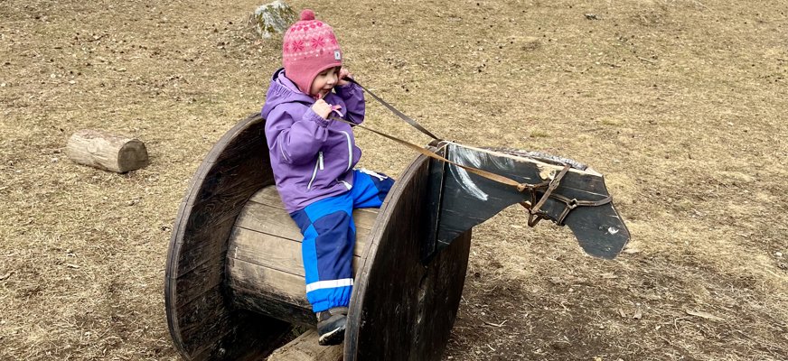 En liten flicka rider på en låtsashäst gjord av en tunna. Hon håller i ett par tyglar och har en rosa mössa, lila jacka och blåa täckbyxor på sig. 