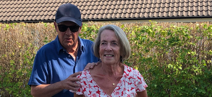 Stellan Andersson i klädd blå pikétröja och solglasögon håller händerna på partnern Elisabeths axlar. Hon är klädd i en röd- och vitmönstrad klänning.