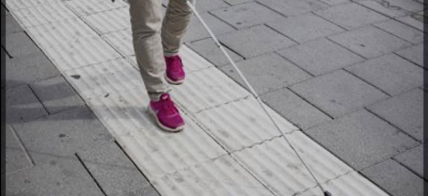 En person med vit käpp går längst med ett ledstråk. Bilden visar benen på personen, klädd i ljusa byxor och röda gympaskor. Vita käppen förs snett framför kroppen och spetsen ligger på det ljusa, räfflade ledstråket som löper längs trottoaren. Runt ledstråket finns släta, rektangulära trottoarplattor i en mörkare grå nyans.