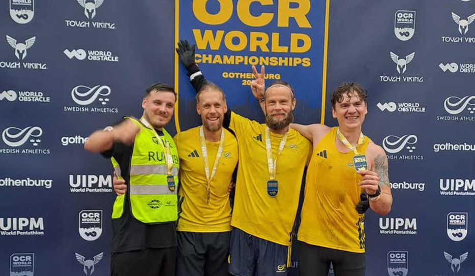Hans tillsammans med sin ledsagare och övriga svenska VM-medaljörer poserar med sina medaljer framför en vägg med texten "OCR World Champions".