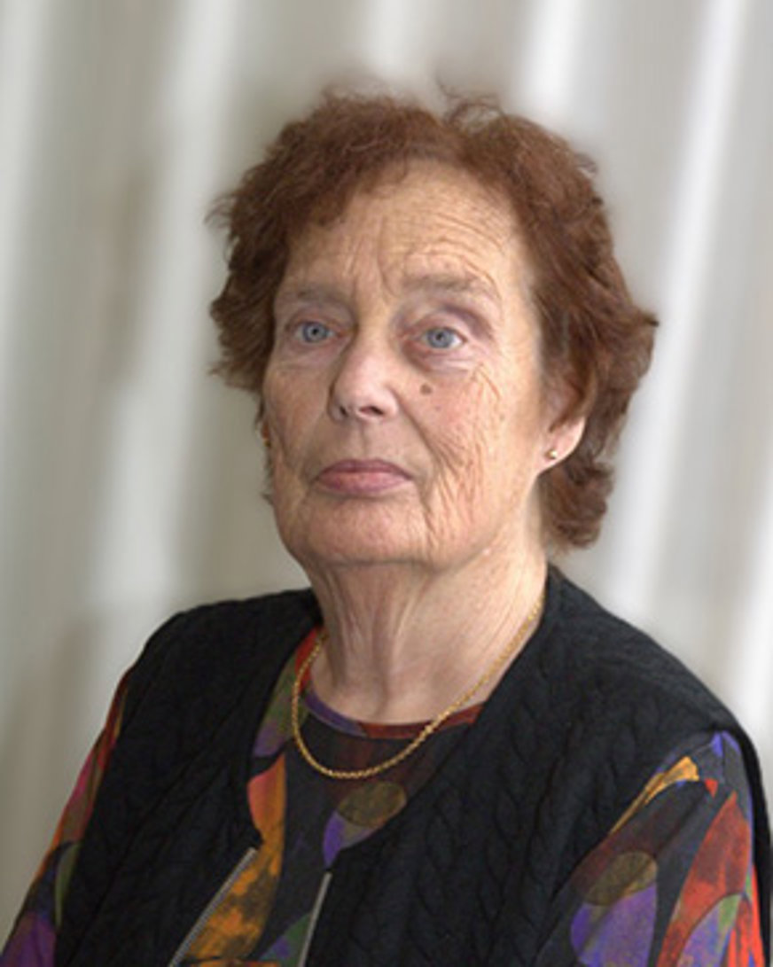 Porträttbild Kristina Adolfsson: Äldre kvinna med rödbrunt hår och färggrann tröja med svart väst över.