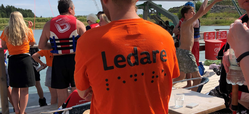Ryggtavlan på någon med orange t-shirt med texten "Ledare" i tryckt svartskrift och punktskrift. I bakgrunden skymtar personer med flytvästar, badkläder, blå sommarhimmel.