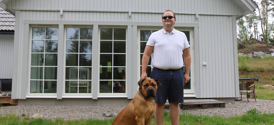 En man står på gräsmattan framför ett vitt trähus med stora glaspartier. Han bär solglasögon, en vit kortärmad pikétröja och mörkblå shorts. Bredvid honom sitter en stor brun hund med svart nos och mörka öron. Hunden sitter nära mannens ben.