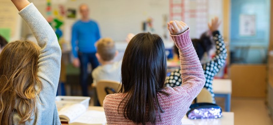 Elever syns bakifrån när de räcker upp handen i ett klassrum. I förgrunden sitter två flickor med långt hår och längst fram vid tavlan står läraren. 