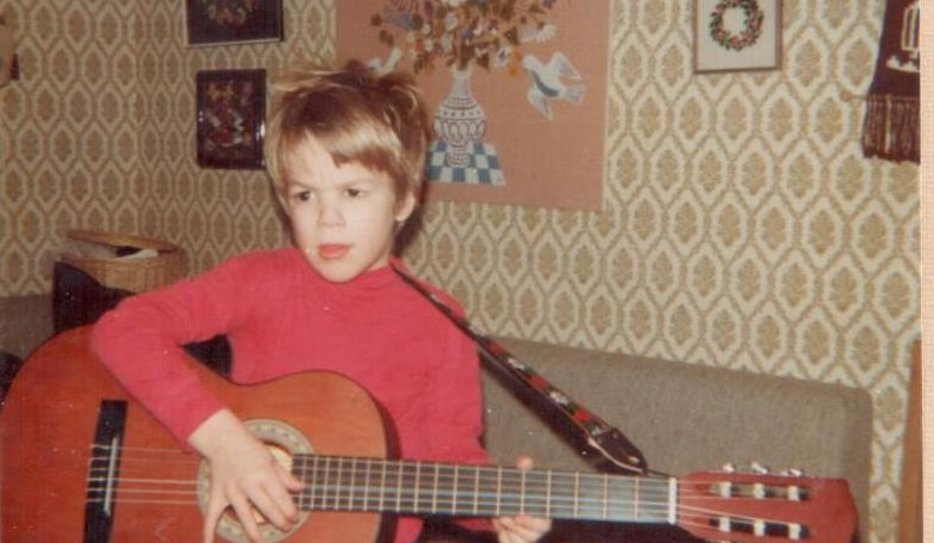 Fredrik som barn, en blond och brunögd pojke i femårsåldern, spelar gitarr. Hans kläder och rummets är inredning är i typisk sjuttiotalstil.