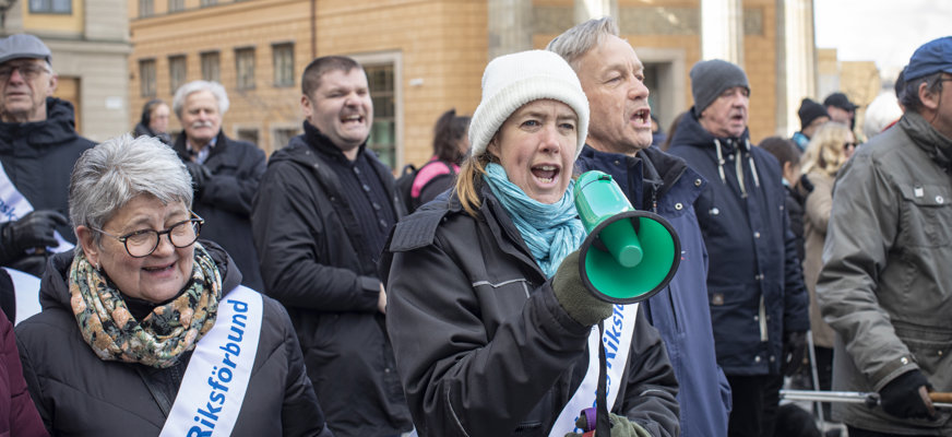 Kvinna med megafon och SRF-banderoll i folkhav, vinterklädsel.