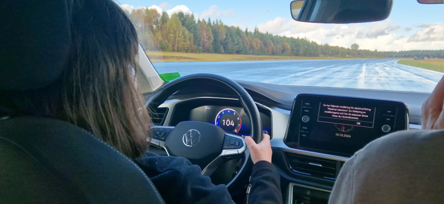 En person sitter och kör med båda händerna på ratten, man ser vägen framför bilen och körledaren som sitter på passagerarsätet.
