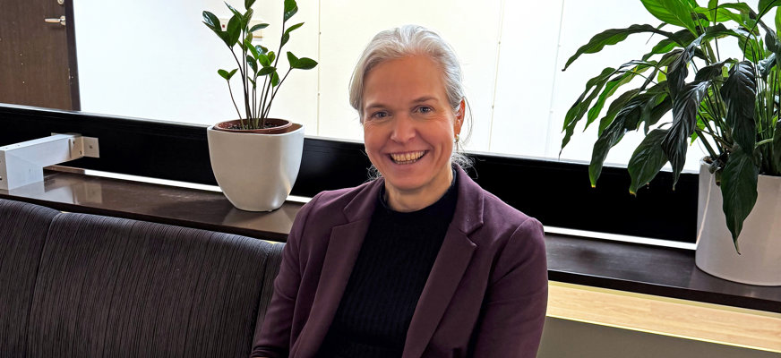 Jenny Ögren sitter i en soffa på SRF:s Rikskansli. Hon har grått, uppsatt hår och en lila kavaj över en svart tröja. Hon ler stort mot kameran.