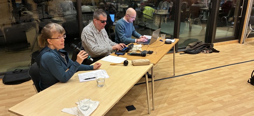 Bilden visar presidiet vid novembermötet. Elin Forsgren talar i mikrofon medan Kjell Emanuelsson skriver på sin punktdisplay och Anton Quarnström skriver på datorn. Mötesdeltagarna speglas i glasväggen bakom presidiet.