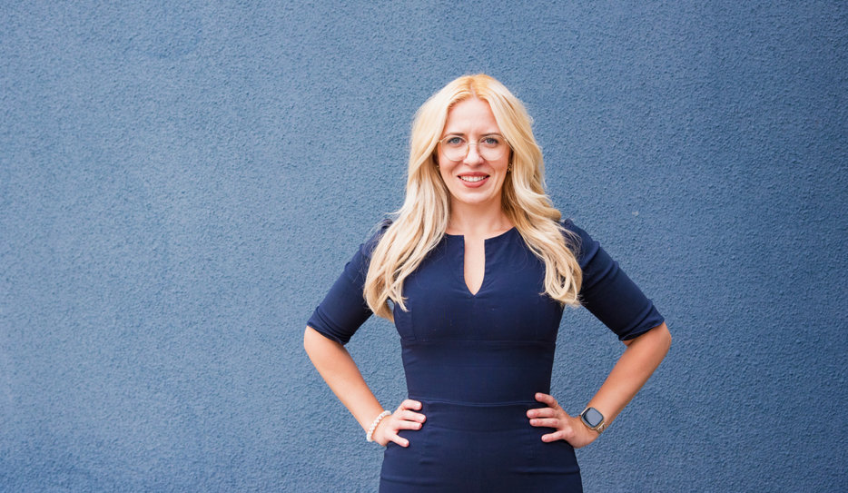 Andreea Badache har blont, långt hår och glasögon med tunna bågar. Hon ler stort, har på sig en blå klänning och står upp med händerna på höfterna.