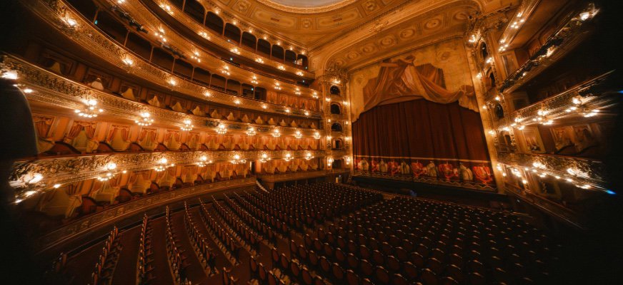 En stor teatersalong med rader av tomma stolar framför en scen med neddragen röd ridå. Salongen är rikt dekorerad i guld och har flera balkonger med lampor som lyser varmt.