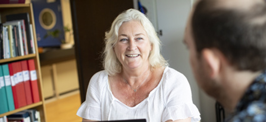 Kvinna sitter framför dator och pratar med man som syns i förgrunden.