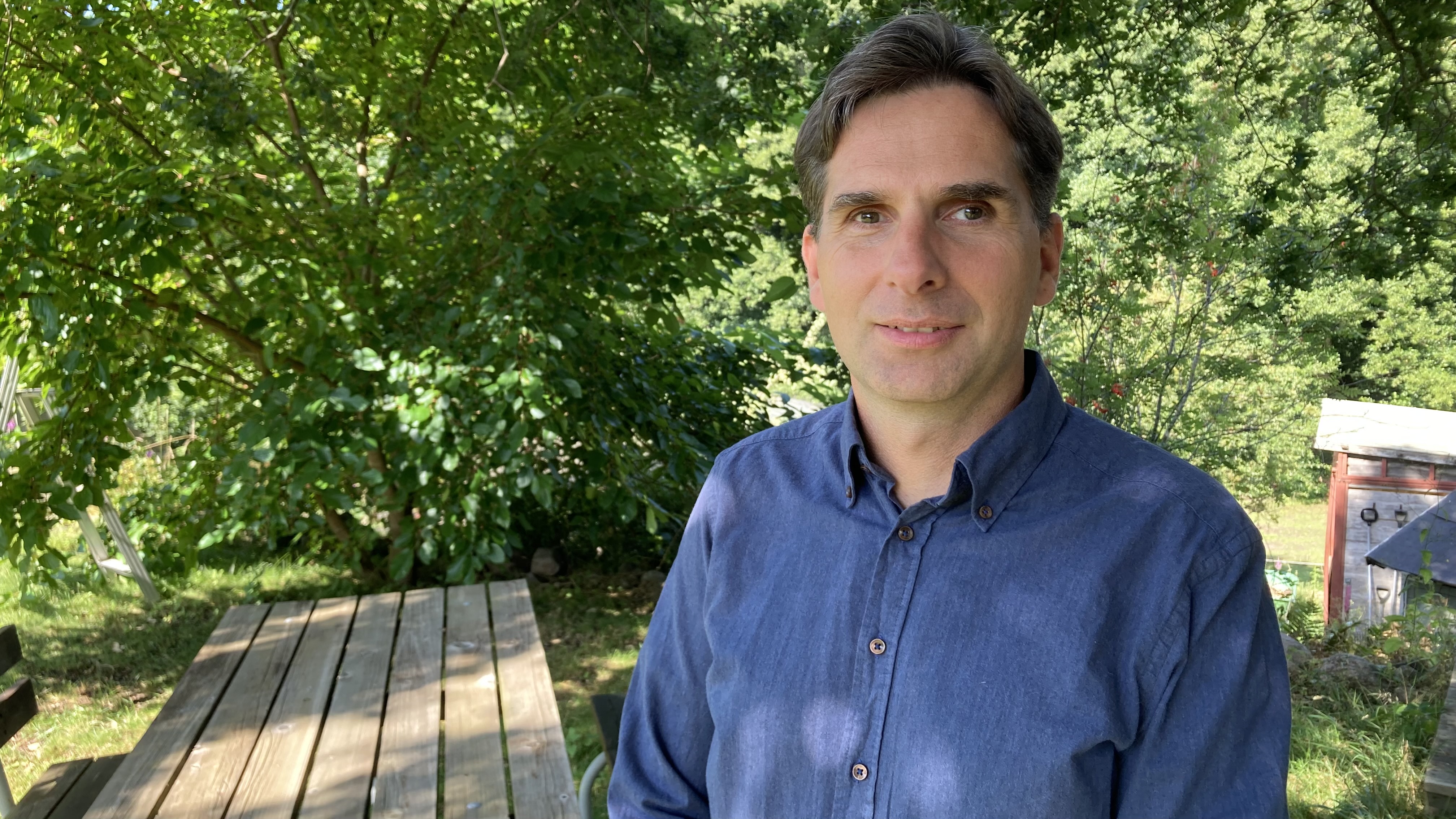 Karl-Otto Rosenquist i blå skjorta i sin trädgård, i bakgrunden syns grönskande buskar och ett utebord i trä.