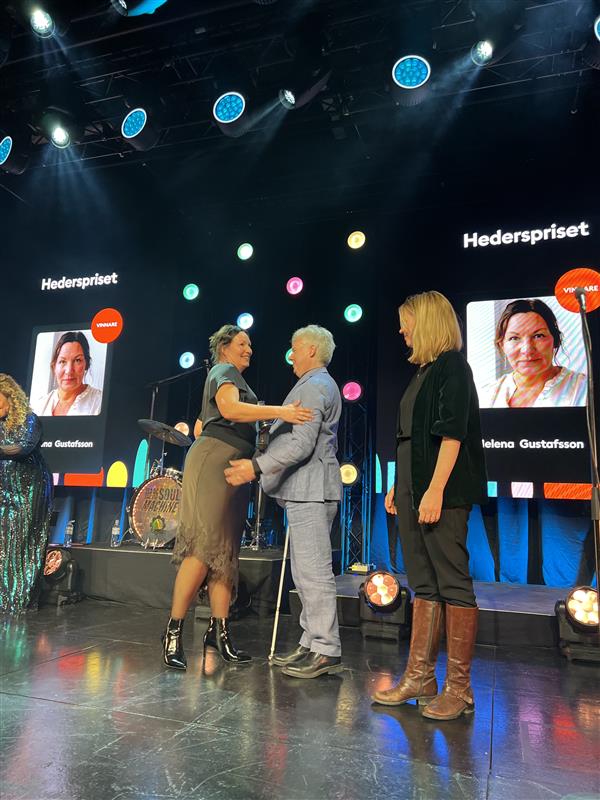 Helena Gustafsson mottar hederspriset av Niklas Mattsson på scenen. De kramas. Bakom dem syns Helenas ansikte på storbildsskärmar.