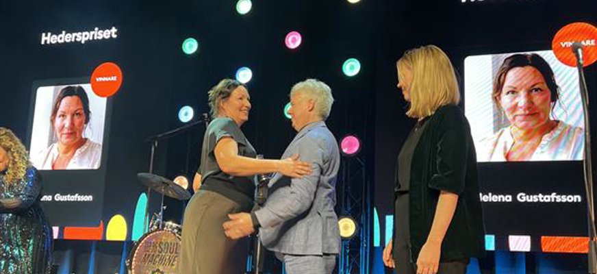 Helena Gustafsson mottar hederspriset av Niklas Mattsson på scenen. De kramas. Bakom dem syns Helenas ansikte på storbildsskärmar.