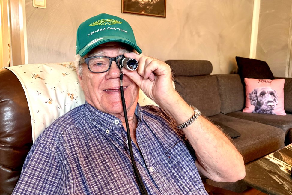 Vit 84-årig man i blåvitrutig kortärmad skjorta och grön keps sitter i fåtölj i sitt vardagsrum. Han håller en liten monokikare framför vänster öga.