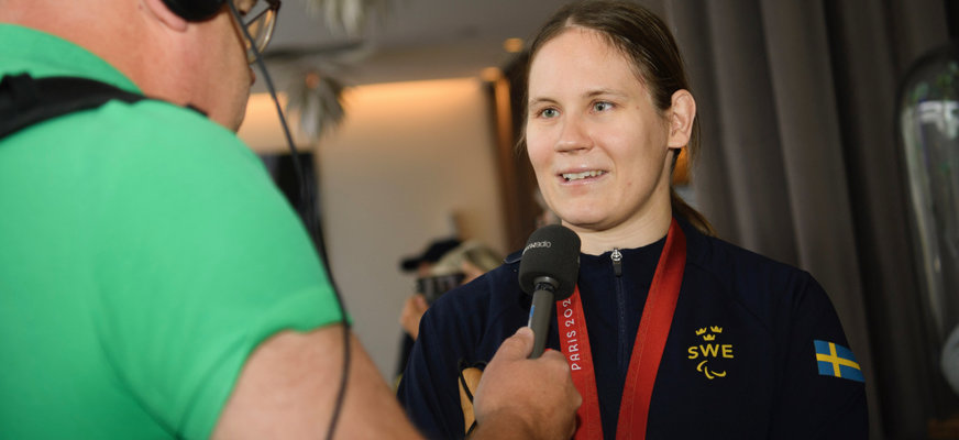 Nicolina intervjuas i samband med Paralympics 2024. I förgrunden syns en man med hörlurar. Han håller upp en mikrofon framför Nicolinas ansikte. Hon ler brett och har på sig en mörk zip-tröja med Sveriges flagga på armen.