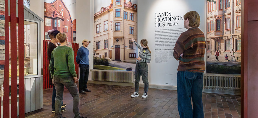 Besökare i Göteborgs stadsmuseums utställning om landshövdingehus. Flera personer tittar på stora väggbilder föreställande landshövdingehus och en väggtext med rubriken “Landshövdingehus 150 år”