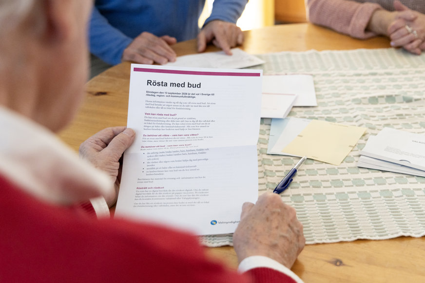 En person sitter vid ett bord och håller upp ett informationsblad med rubriken ”Rösta med bud”. På bordet ligger fler papper, kuvert och en penna. Ytterligare personer sitter på andra sidan bordet.