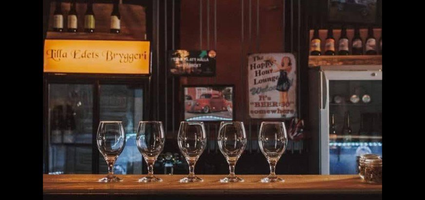 Fem tomma glas står uppradade på en bardisk av trä. I bakgrunden syns en skylt med texten "Lilla Edets Bryggeri" och ett antal ölflaskor står på två hyllor. På väggen finns en affisch med en tecknad kvinna och texten "The Happy Hour Lounge".