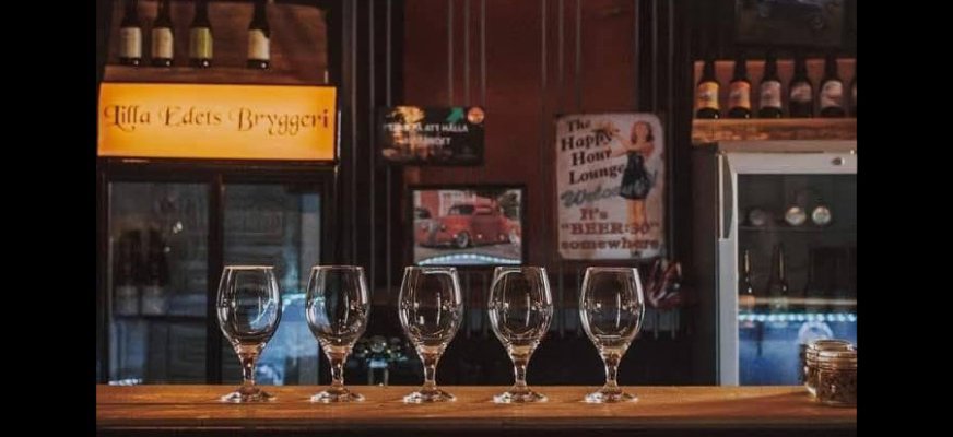 Fem tomma glas står uppradade på en bardisk av trä. I bakgrunden syns en skylt med texten "Lilla Edets Bryggeri" och ett antal ölflaskor står på två hyllor. På väggen finns en affisch med en tecknad kvinna och texten "The Happy Hour Lounge".