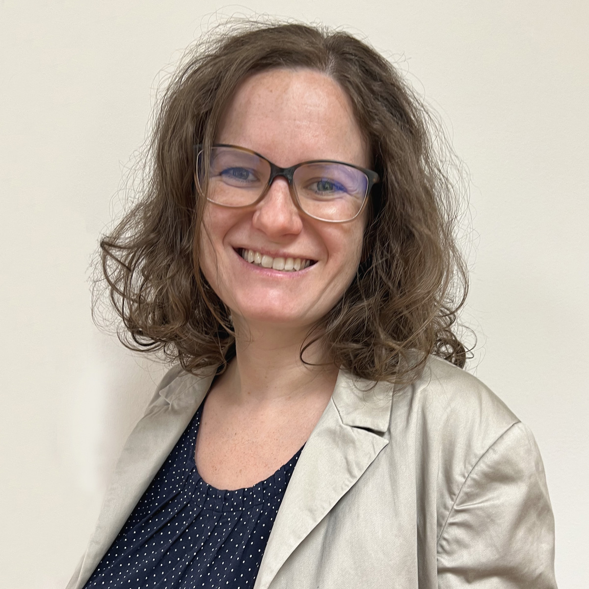 Leende kvinna i 30-årsåldern, lockigt brunt axellångt hår, bruna glasögon, beige kavaj, prickig blus.