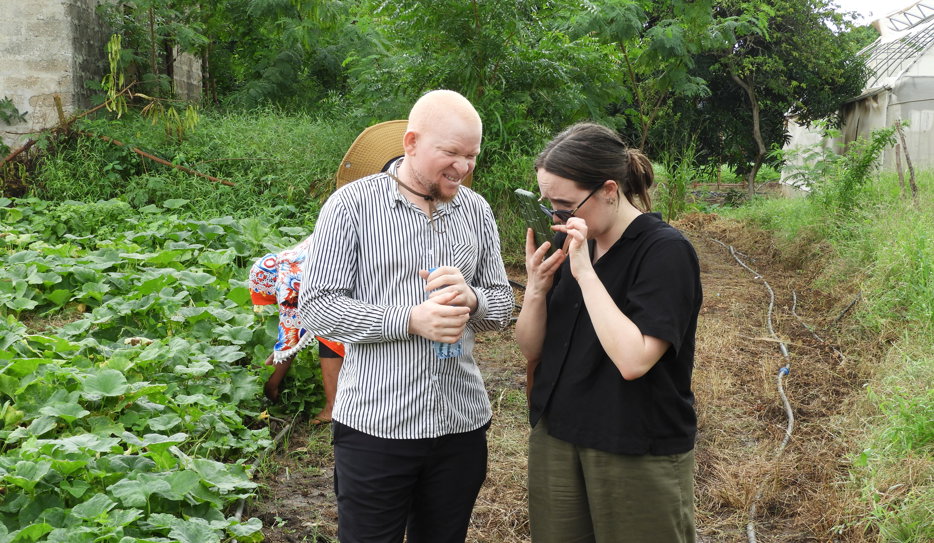 Två personer står i ett grönsaksland och kollar på en telefon tillsammans. Den ena personen är svensk och kollar på sin mobilskärm med förstoring. Den andra personen är Tanzanian och har albinism.