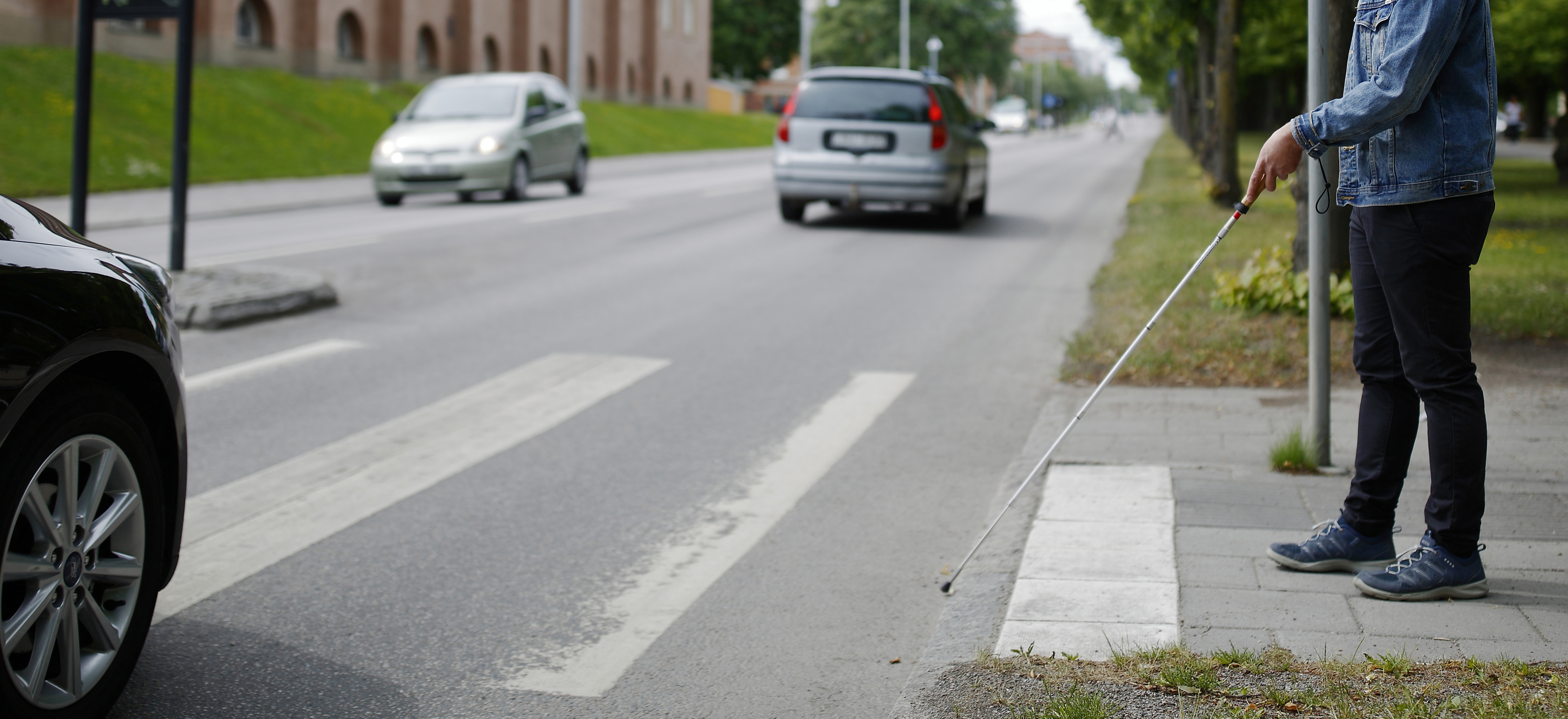 En man med vit käpp som väntar vid ett övergångsställe för att kunna korsa en bilväg.