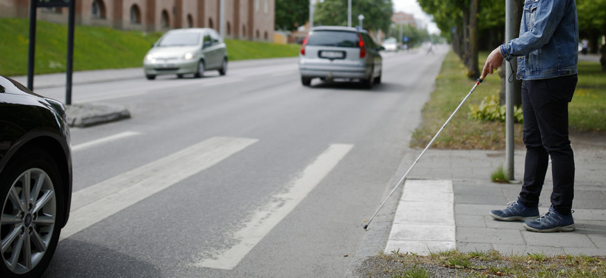 En man med vit käpp som väntar vid ett övergångsställe för att kunna korsa en bilväg.